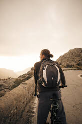 Young male backpacker with bicycle looking at mountains against sky - MASF40759