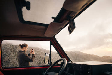 Ein junger Mann bewundert die atemberaubende Berglandschaft aus dem Fenster eines Lieferwagens und hält sie mit seinem Smartphone fest - MASF40787