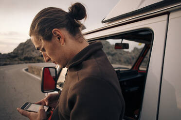 Ein Mann mit einem eleganten Dutt genießt seinen Urlaub und schreibt eine SMS auf seinem Handy, während im Hintergrund ein Lieferwagen parkt - MASF40789