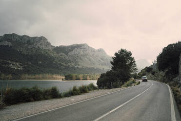 Ein Van fährt durch eine malerische Landschaft, flankiert von einem Fluss und hoch aufragenden Bergen, unter einem heiteren blauen Himmel - MASF40793
