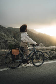 Eine Frau bewundert den atemberaubenden Blick auf den Sonnenuntergang, während sie mit ihrem Fahrrad auf einer Bergstraße steht - MASF40804