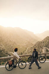 Multiracial male and female friends looking at mountains while wheeling bicycles on road - MASF40810