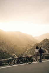 Friends looking at mountains while standing roadside with bicycles at sunset on vacation - MASF40811