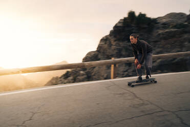 Ein geübter Skateboarder fährt bei Sonnenuntergang anmutig eine Straße entlang und manövriert sich mühelos um das Geländer herum - MASF40819