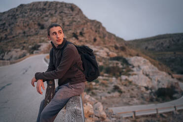 Ein junger Mann bewundert die atemberaubende Bergkulisse, während er auf einem Geländer sitzt und den Sonnenuntergang beobachtet - MASF40822