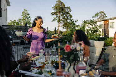 Eine Frau serviert ihren Freunden auf einer Gartenparty fröhlich das Essen, während sie gemeinsam an einem Esstisch sitzen - MASF40875