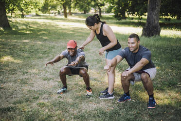A female coach assists men in improving their squat form on the grassy terrain of a park - MASF40979
