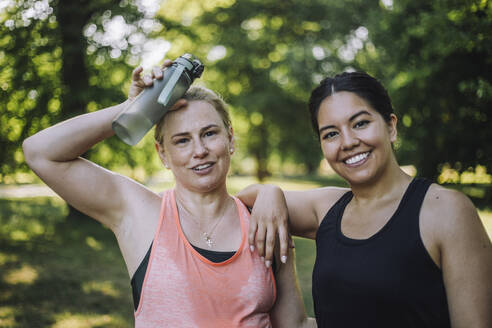 Portrait of smiling woman with exhausted female friend holding water bottle at park - MASF40990