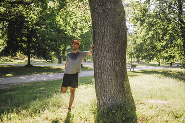 Man doing stretching while standing near tree at park on sunny day - MASF40992