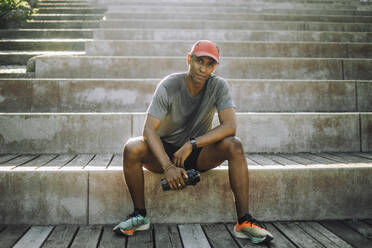 Portrait of exhausted man holding shaker bottle while sitting on steps - MASF40996