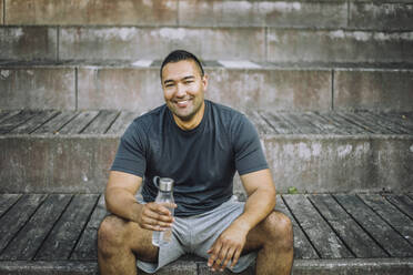 Portrait of smiling man holding water bottle while sitting on steps - MASF41017