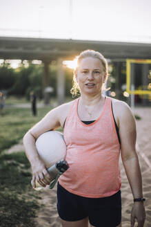 Sportliche Frau, ausgerüstet mit Wasserflasche und Volleyball, bereit für einen vergnüglichen Tag am Strand - MASF41072