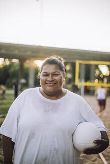 Eine fröhliche Plus-Size-Dame posiert für ein Porträt, während sie einen Volleyball hält und ein weißes T-Shirt trägt - MASF41074