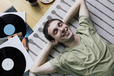 High angle view of happy man lying down near vinyl records on carpet at home - MASF41094