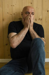 A worried man in his middle age sitting by a wooden wall at home, with his hands covering his mouth in anxiety - MASF41109