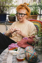 An elderly woman finds joy in knitting a warm sweater while taking a break on her balcony - MASF41173