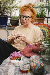 Eine ältere Frau strickt mit Freude einen warmen Pullover, während sie auf ihrem Balkon eine Pause einlegt - MASF41173