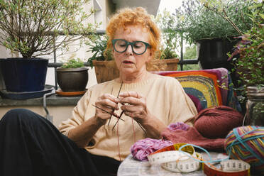 An elderly woman indulges in her passion for knitting, crafting a warm and comfortable sweater on her balcony - MASF41174