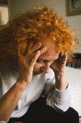 Eine ältere Frau mit roten Haaren sitzt auf dem Bett und stützt ihren Kopf in die Hände, während sie verzweifelt und belastet aussieht - MASF41182