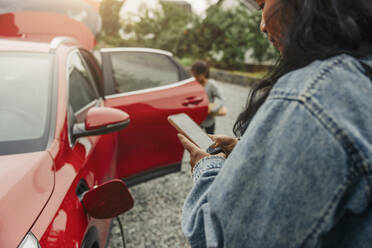 A woman monitoring her electric car's charging status on her phone while standing next to the vehicle - MASF41194