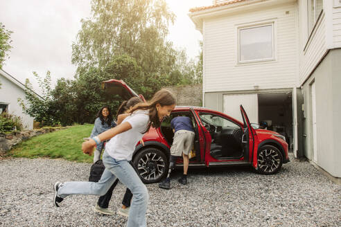 Zwei fröhliche junge Mädchen vergnügen sich neben dem Elektroauto ihrer Familie, das vor ihrem Haus geparkt ist - MASF41199