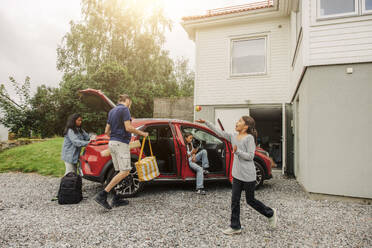 A young girl happily plays with a ball as her family prepares to leave in their electric car from their home - MASF41200
