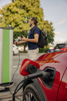 Ein Mann lädt sein Elektroauto an einer Station mit eingestecktem Ladekabel auf, während er im Hintergrund steht - MASF41219