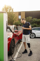 Ein Mann mit einer Beinprothese fördert den nachhaltigen Transport, indem er sein Elektroauto an einer Ladestation auflädt - MASF41221