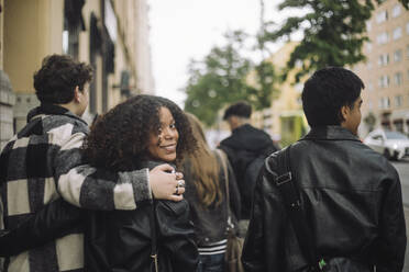 Boy with arm around female friend looking over shoulder while walking at street - MASF41251