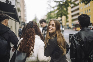 Portrait of smiling girl looking over shoulder while walking with friends at street - MASF41255