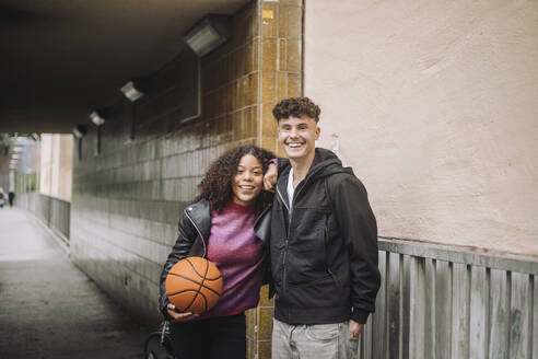 Portrait of smiling girl holding basketball while standing with male friend near underpass - MASF41279