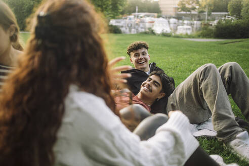 Four friends enjoying a sunny day in the park, chatting and laughing while lounging on the grass - MASF41318
