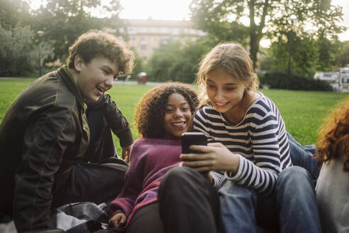 Drei Freunde im Park, ein Mädchen im Teenageralter, das sein Smartphone mit einem männlichen und einem weiblichen Freund teilt - MASF41320
