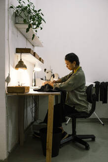 Eine Näherin arbeitet an einer Nähmaschine, während sie in einer Werkstatt sitzt und Kleidung näht (Seitenansicht) - MASF41373