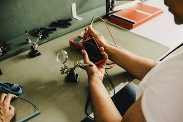 A male technician is seen examining a smart phone using a multimeter from a high angle view at a repair shop - MASF41401