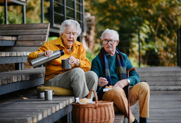 Ein fröhliches älteres Ehepaar genießt ein Picknick am See und entspannt sich nach einem erfrischenden Herbstspaziergang. Die Ehefrau gießt Tee aus einer Thermoskanne ein - HPIF31667