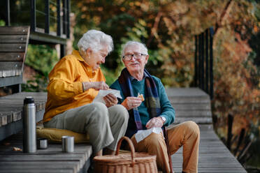 Fröhliches älteres Paar, das nach einem erfrischenden Spaziergang in gemütlicher Herbstkleidung ein Picknick am See genießt - HPIF31668