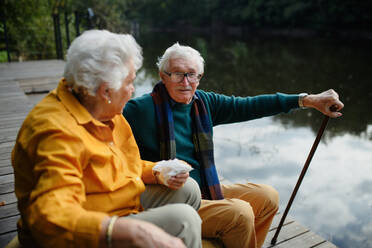 Ein lächelndes älteres Ehepaar ruht sich am See aus und genießt die frische Herbstluft in ihren gemütlichen Kleidern - HPIF31675
