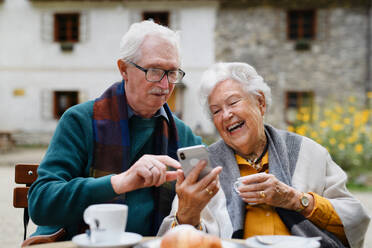 Elderly couple enjoying a relaxing break at a park cafe, sipping coffee and browsing on their smartphone - HPIF31694