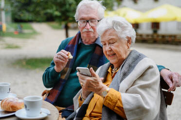 Elderly couple enjoying a break at a park cafe, sipping coffee and browsing their phone after a refreshing walk - HPIF31695