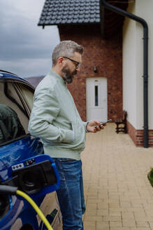 A mature man patiently waits for his new electric car to charge up. - HPIF31743