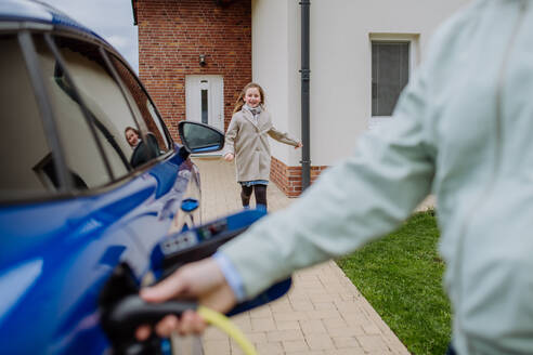 A man charges his new electric car while his daughter runs to him, holding a power supply cable in close-up - HPIF31749