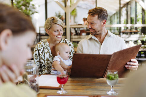 Eine dreiköpfige Familie, darunter ein Baby, sieht sich die Speisekarte an und wählt ihr Essen während eines gemütlichen Abendessens in einem Restaurant aus - HPIF31772