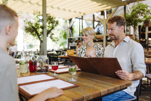 Eine dreiköpfige Familie, darunter ein Baby, sieht sich die Speisekarte in einem Restaurant an und entscheidet sich für ihre Speisen und Getränke für ein gemütliches Abendessen - HPIF31774