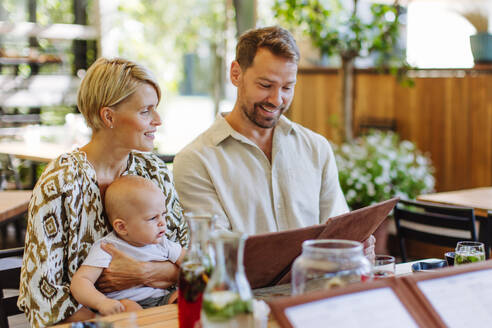 Eine dreiköpfige Familie isst in einem Restaurant zu Mittag, wobei die Mutter ihr Baby im Arm hält, während sie die Speisekarte studiert - HPIF31777