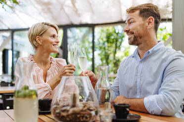 Portrait of beautiful couple in a restaurant, on a romantic date. Husband and wife are clinking champagne glasses, making a toast at restaurant patio. - HPIF31796