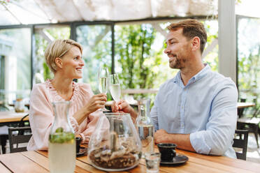 Portrait of beautiful couple in a restaurant, on a romantic date. Husband and wife are clinking champagne glasses, making a toast at restaurant patio. - HPIF31797