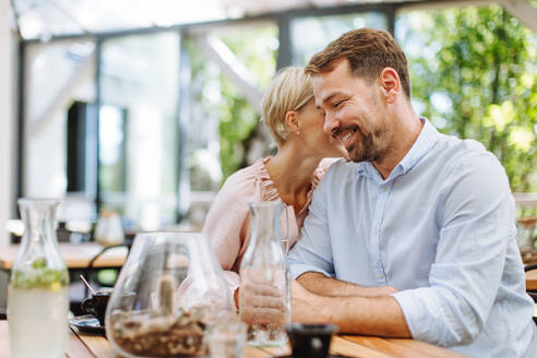 Ein verliebtes Paar genießt einen romantischen Moment beim Abendessen auf einer gemütlichen Restaurantterrasse, wobei die Frau ihrem Mann süße Worte zuflüstert - HPIF31809