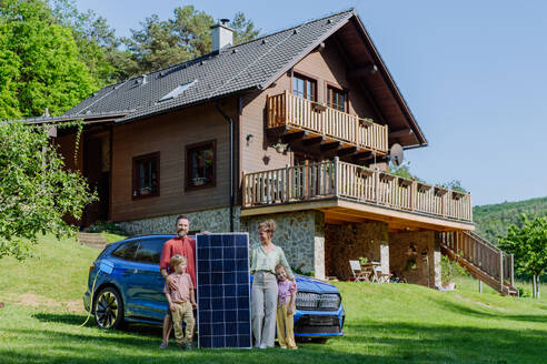 A joyful family stands proudly next to their solar panel while charging their electric car - HPIF31819