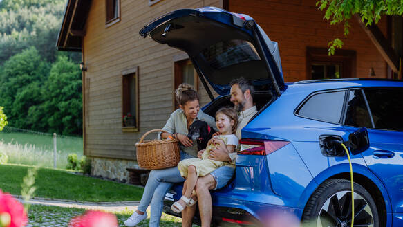 Eine fröhliche Familie wartet darauf, dass ihr Elektroauto aufgeladen wird, bevor sie sich bequem aus dem Kofferraum auf ein Picknickabenteuer begibt - HPIF31829